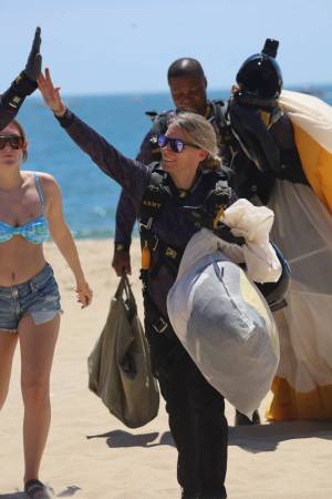 Group of skydivers on a sunny beach celebrating a successful jump, with parachute gear and ocean behind them.